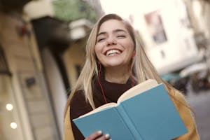 Smiling woman enjoying a book with earbuds in an urban outdoor setting.